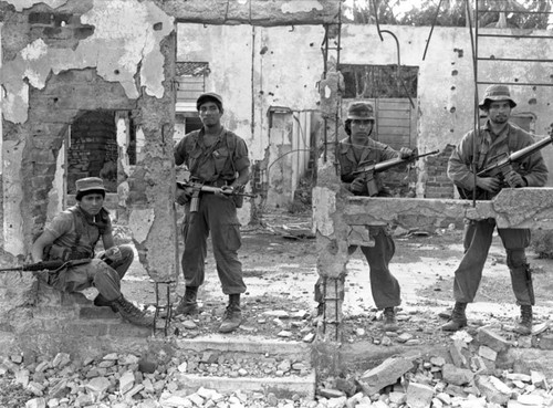 An armed squad stands in front of destroyed building, Perquín, 1983