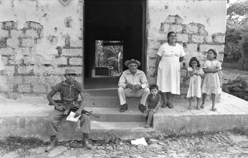 A soldier sits on the steps of a house with residents standing next to him, Perquín, 1983