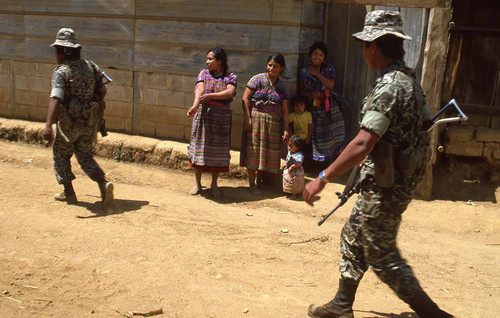 Two soldiers walk past Mayan women and children, Zaragoza, 1982