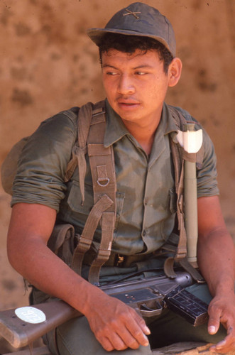 Soldier sitting with weapon on his legs, San Antonio de los Ranchos, Chalatenango, El Salvador, 1981