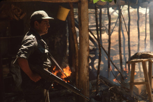 Soldier standing next to a burning farmhouse near Suchitoto, El Salvador, 1981