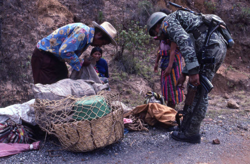 A soldier inspecting a Mayan man's basket in search of subversive materials, Chichicastenango, 1982