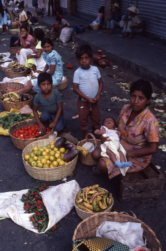 A woman holding a baby and selling produce at the market, Chiquimula, 1982