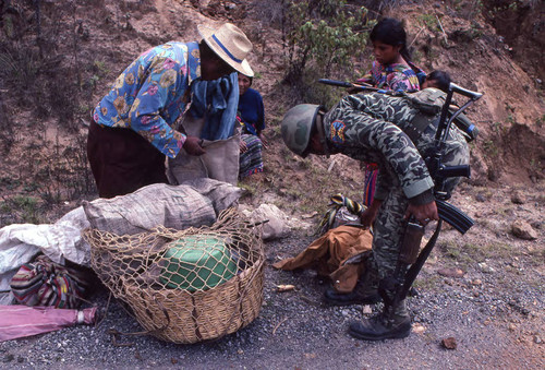 A soldier inspecting a Mayan man's basket in search of subversive materials, Chichicastenango, 1982