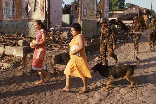 Two women and a dog walk ahead of soldiers, San Agustín, Usulután, 1983