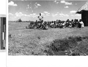 Group of boys sitting in rows on the ground outside a hut, Africa, July 1950