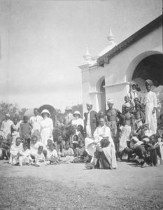 Hospitalet i Tirukoilur, Sydindien. Udenfor Dispensariet, marts 1915. Med vognen som baggrund ses 1. assistent Mr. Amos Lucas og sygeplejerske Agathe Poulsen. Nærmere mod bygningen ses jeg (Dr. Chr. Frimodt-Møller?) og min kone. Mod den hvide mur ses 2. assistent, Mr. Radnam Isaac med den sorte trøje. Midt i den store portåbning ses 3. assistent, Mr. Milton Thomas. Foran Mr. Radnam sidder en Brahmin