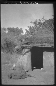 African woman lying on the ground in front of a hut, Mozambique, ca. 1933-1939