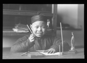 Smiling boy seating at a table writing, China, ca. 1918-1938