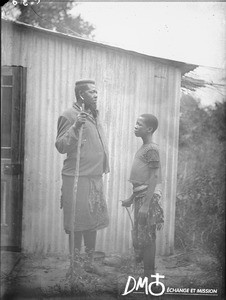 African man and boy standing in front of a corrugated iron building, Antioka, Mozambique, ca. 1901-1915