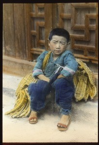 Young boy seated outside of a building, carrying bundles of sandals, China, ca.1917-1923