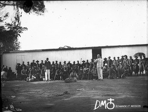 African men sitting and standing in rows in front of a corrugated iron building, Maputo, Mozambique, ca. 1901-1915