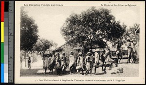 People bearing burdens as they walk outside a small stone building, India, ca.1920-1940