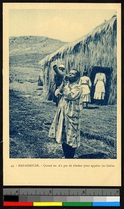 Man blowing hollowed horn outside a thatched building, Madagascar, ca.1920-1940