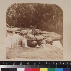 Man seated on rock by waterfall in forest, Madagascar. ca. 1865-1885