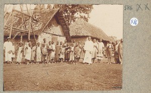 African schoolchildren with teacher(?) in front of a belfry(?) and rectangular buildings with plant fiber roofs, ca.1900-1914