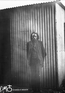 African man standing in front of a corrugated iron building, Antioka, Mozambique, ca. 1901-1915