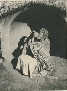 A woman drinking some beer in a calabash, in the entrance of her hut