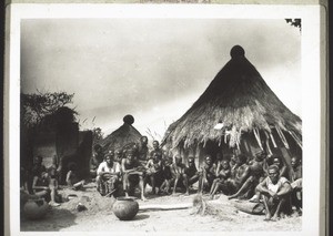 People listening to open-air preaching in Mbembe (Grassfields). Chief with goitre, next to him the teacher, a christian, and on the right someone is working at turning bark into cloth