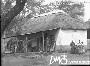 Arnold Borel and African people standing in front of a building with a thatched roof, South Africa, ca. 1896-1911