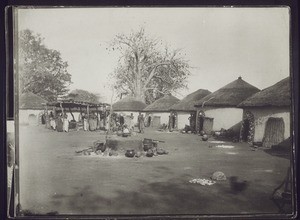 Dagomba compound with fireplace in the centre, and a baobab in the background. The black points on the baobab are fruits