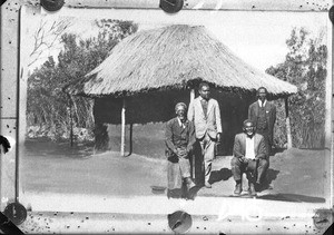 African men sitting and standing in front of a building with a thatched roof