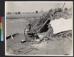 Woman preparing food outside a grass hut on an arid plain, India, ca.1919-1943