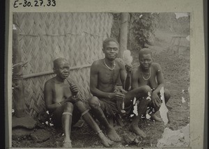 Man spinning, with his wives, behind them his hut