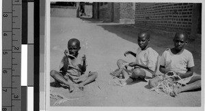 Three boys sitting outside making rope, Africa, March 1949