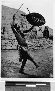 Man dancing by a stone wall with two sticks and a shield, Africa, 1939