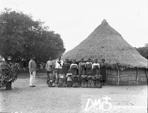 African teacher and students in front of a building with a thatched roof, Antioka, Mozambique, 1904