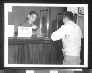 "George Haramoto (Nisei), teller at the branch of the California Bank in Los Angeles, located in the Japanese quarter of the city, shown taking care of a customer."--caption on photograph