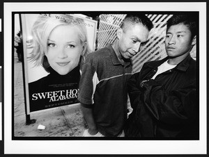 Two young men of South American origin standing outside, with a poster of a film star behind, Mission District, San Francisco, California, 2002