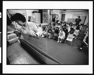 Woman holds cue card with back turned to audience of children, Oriental (Los Angeles), 1996