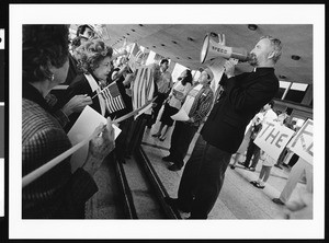 Fr. David O'Connell speaking to group through megaphone in front of protesters holding signs, St. Thomas, Los Angeles, 1996