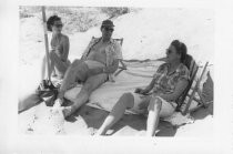 Two women and one man under umbrella at beach, 1954