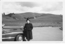 Woman standing next to automobile with hills in background, unknown