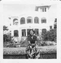 Woman sitting on outside stone fence with house in background, unknown