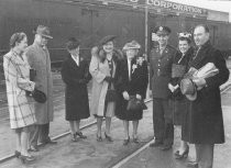 Five women and three men standing outside in front of passenger train, unknown