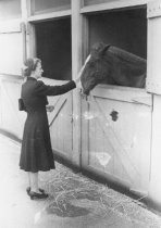Woman standing next to horse stable touching horse's nose, unknown