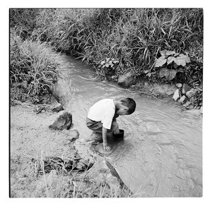 Boy wearing boots steps crouches down into a stream, Korea