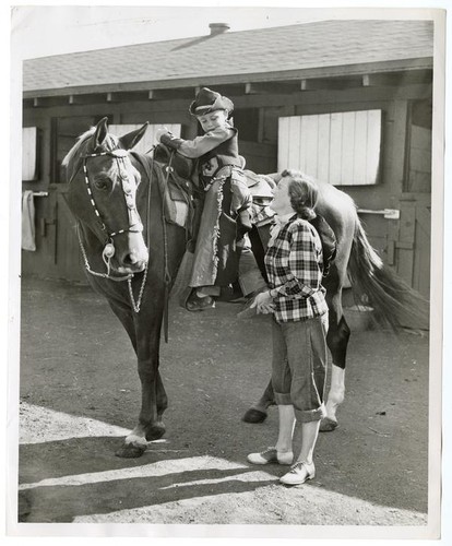 Young boy, Tim Turner, dressed as a cowboy on horseback with a woman, Mrs. John Zafferano, looking on