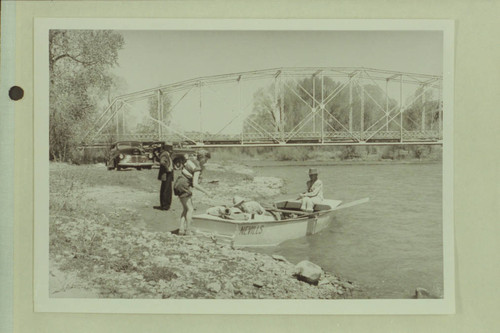 Final stowage of the punt before start of the Dolores River traverse. Bridge below Dolores, Colorado. Becky Walker stands near the boat. Marston is stowing dunnage under the deck and Margaret Marston sits on the boat