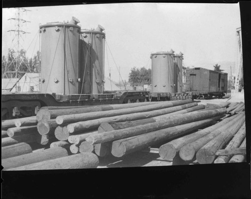 Storage yard with poles on ground and large electric transformers being delivered on railroad flatcars