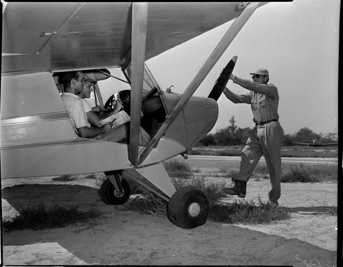 Two people sitting in cockpit of Piper Cub airplane while man turns the prop to start the engine by hand