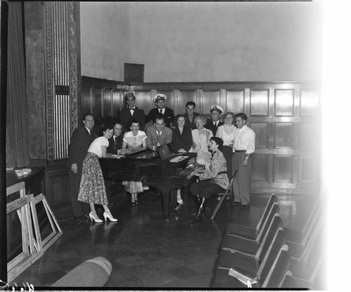 Group gathered around a lady playing a grand piano in the corner of an auditorium near a stage