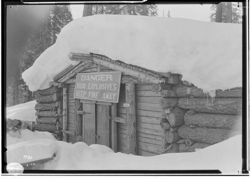 Log constructed Powder Magazine in the snow at Camp 60 with sign over doorway "Danger High Explosives Keep Fire Away"]