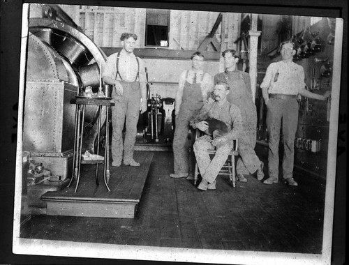 Five men (one holding a dog) standing next to the switchboard and generators in Mill Creek #1 Hydro Plant