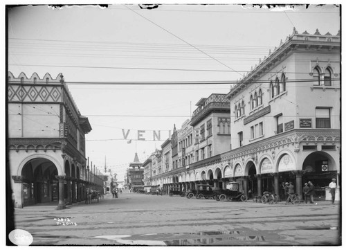 An area view of Windward Avenue in Venice, California, [showing a Turkish Bath House, theaters, an apartment house and automobiles common to 1914 parked along the street. There is a large sign at the intersection that reads: VENICE. People are socializing along the street and there is a horse