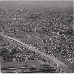 Aerial view, looking north from around East Creek Slough, of the Nimitz Freeway being constructed through the Kennedy Tract district of Oakland, California. Montgomery Ward distribution center, Lake Merritt, and El Cerrito Hill in view